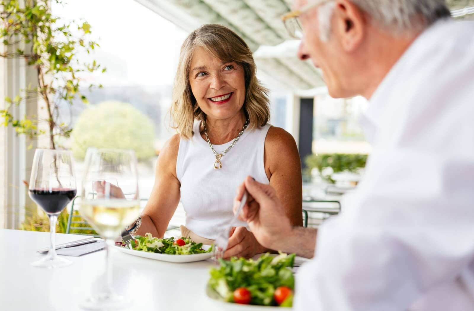 Older person dining at a restaurant table smiling at their companion while enjoying a fresh salad and glass of white wine