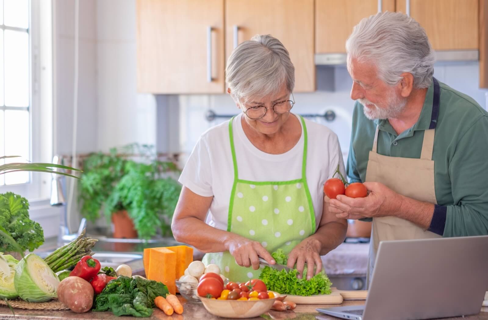 Two older adults preparing fresh vegetables together in a kitchen surrounded by leafy greens tomatoes and seasonal produce