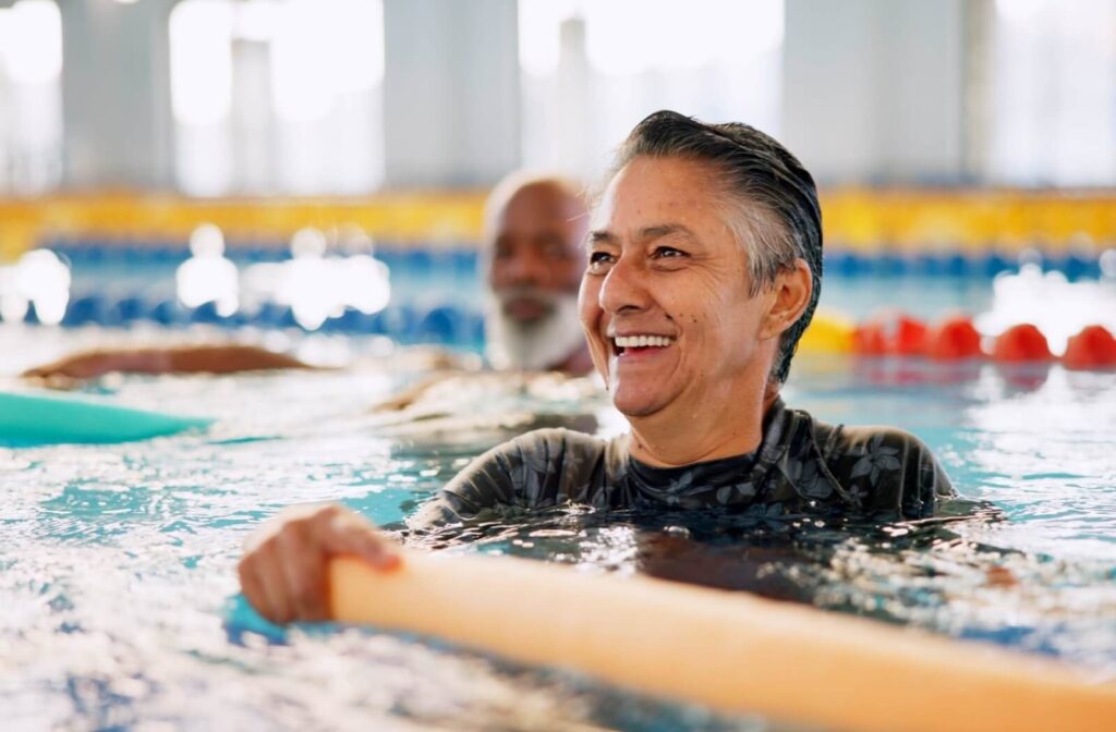 An older adult smiles while swimming in a pool with a pool noodle to strengthen their balance