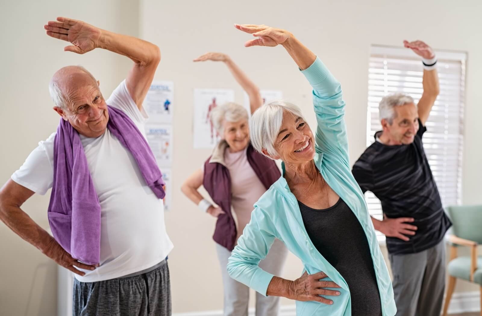 4 older adults stretch their arms overhead and bend to one side while smiling during a balance fitness class in senior living